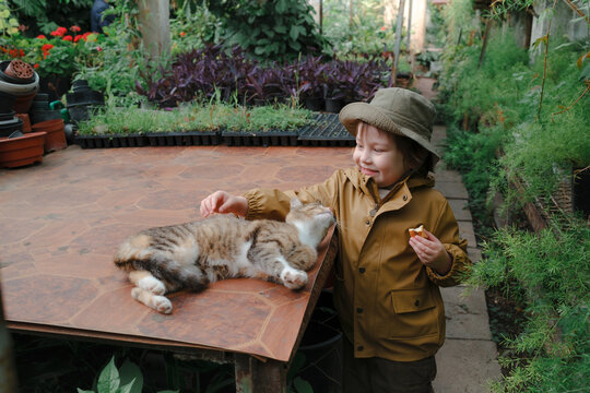 Child exploring greenhouse filled with plants, cat sitting on a table
