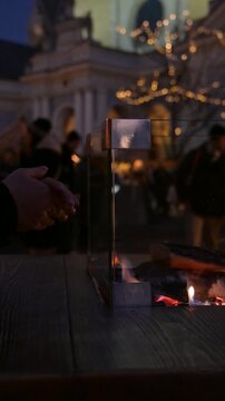 Hands Warming by Fire at Christmas Market in Winter Evening with People Walking in Background