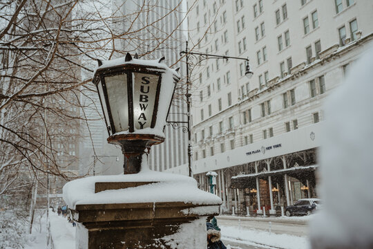 New York, USA, 25 December 2025: Vintage subway lamp post covered in snow near Plaza Hotel. Close up of old subway light fixture with snow against blurred luxury hotel background.