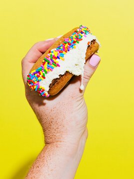 Hand holding an ice cream sandwich with colorful sprinkles against a bright yellow background, showcasing a delicious dessert treat in a close-up view