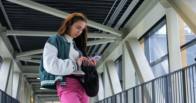 Young woman checking smartphone on walkway, varsity jacket and pink pants, standing by metal railing in glass corridor, scrolling messages with focused expression, urban commuter mood and soft