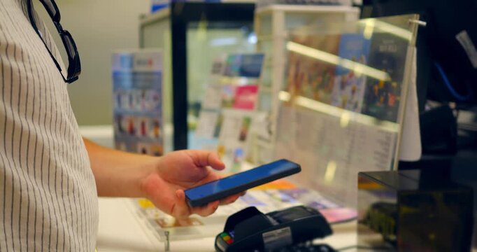 Person using smartphone for mobile payment at store checkout, hands holding phone near card reader and receipt display on payment terminal in retail setting, ready for contactless transaction