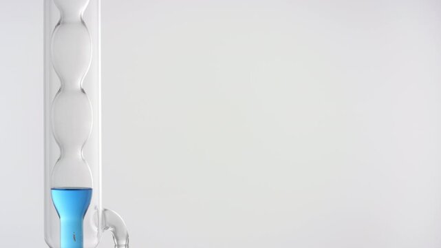 Close-up of clear blue liquid filling up a vertically mounted bulb lab condenser on the left side of the frame, right side for copy space on grey | Methylene blue solution cosmetic extract concept