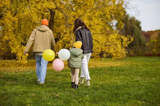 Family with parents and child walking in autumn. Holding balloons, they stroll together in a sunny park, enjoying outdoor time, fresh air, and nature. Concept of family bonding outdoors.