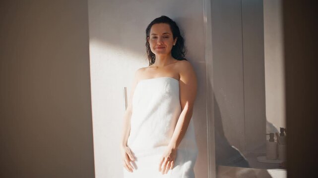 Relaxed woman wrapped towel leaning wall sunlit bathroom after shower portrait