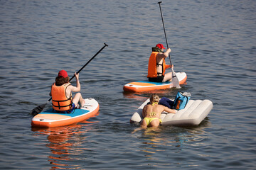 Obraz premium Sup surfing, two guys and girl sailing on a boards and inflatable mattress in water. Stand up paddle boarding in summer