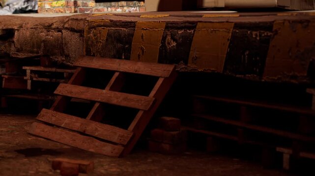 Bags are stacked on one side of a location with wooden pallets arranged nearby. Light comes in from above, highlighting the setup. This scene shows a storage or work area.