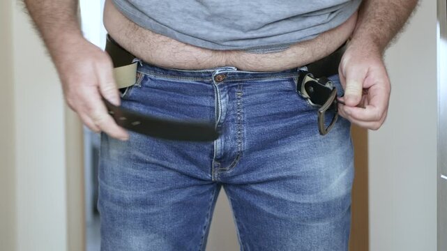 Overweight Man Struggling to Fasten Belt on Jeans Closeup. Man with Big Belly Trying to Buckle Leather Belt.