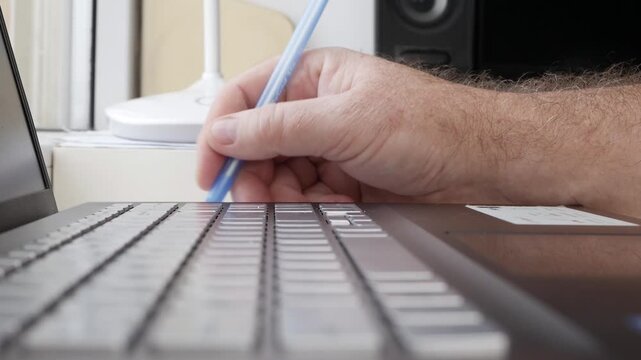 Hand Holding Blue Pen Writing on Laptop Keyboard. Typing on Laptop Keyboard Closeup Detail.