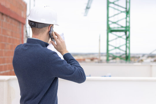 Engineer wearing a hard hat. Standing outdoors at a busy construction site. Communicating on a smartphone while reviewing blueprints