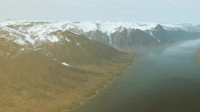 Mountains covered in snow rise above a river that flows between their bases. The landscape shows brown land patches near the water. The scene looks cold and untouched.