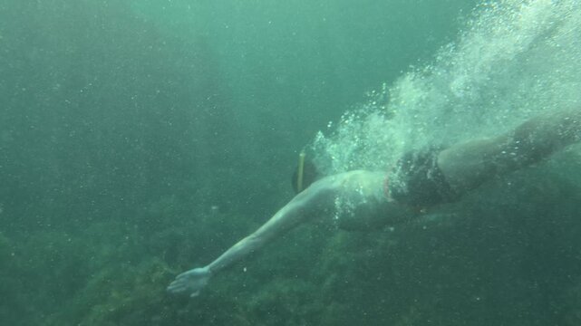 Snorkeling man diving into tropical mediterranean coastal waters from a rocky beach. Underwater split-level tracking shot transitions to deep blue submersion.