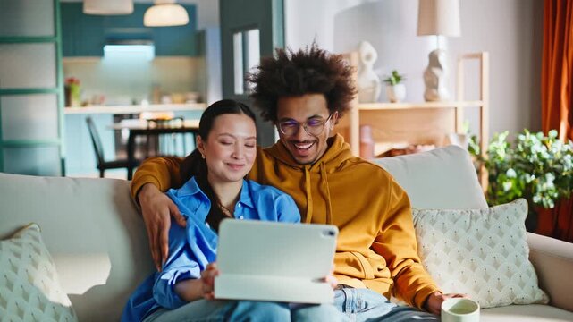 Mixed race couple browsing tablet on sofa. Happy pair relaxing weekend home.