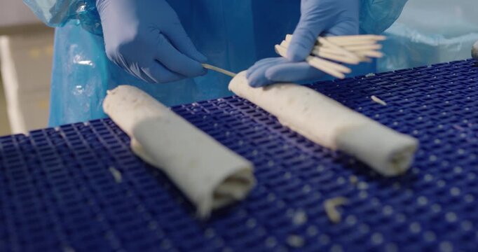 Gloved hands inserting wooden skewers into rolled meat rolls
