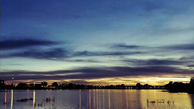 The evening sky glows with warm orange and pink hues, mirrored perfectly on the still water's surface. Distant land silhouettes frame this tranquil, colorful twilight scene.