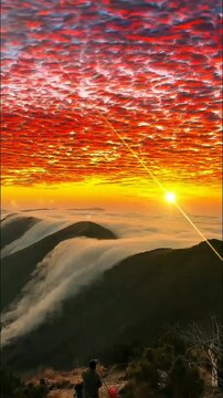Fiery red clouds ignite the dawn sky above majestic mountain peaks. A river of mist cascades through valleys, as observers capture this breathtaking natural spectacle.