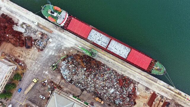 A cargo ship is at the dock beside a large pile of scrap metal and materials. Heavy equipment moves around the area, showing busy port operations during the day.