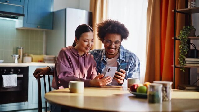 Young couple comparing smartphones at home kitchen. Cheerful pair laughing