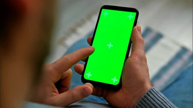 A hand holds a modern smartphone featuring a vivid green screen with tracking markers, ideal for digital overlay. The device is poised for interactive content display.