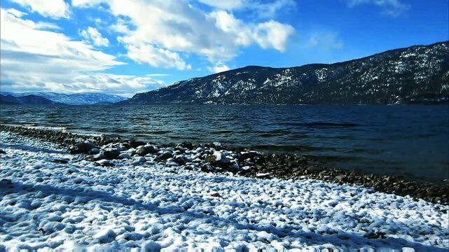 A winter landscape features a snow-covered shore, dark rippling water, and majestic snow-capped mountains under a bright, cloudy sky. A serene, cold beauty.