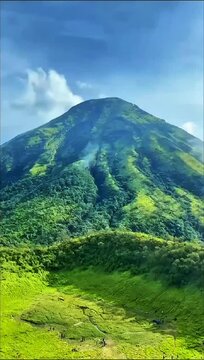 A majestic, verdant mountain dominates a vibrant landscape under a blue sky. Lush slopes meet a valley floor, where small figures explore winding trails.