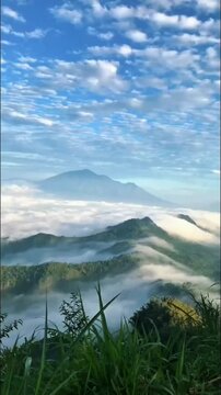 Vibrant grass frames a majestic mountain landscape. Rolling hills are enveloped in ethereal clouds, with distant peaks under a bright, blue, fluffy sky.