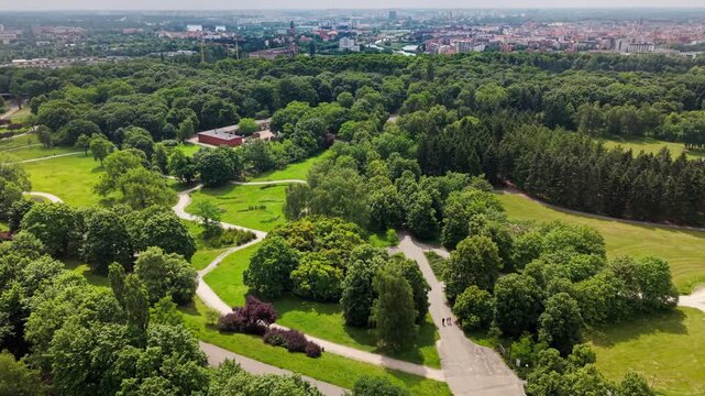 aerial perspective capturing a sprawling urban parkland with large green spaces, lush forests, winding pathways, and a group of people gathered, drone view