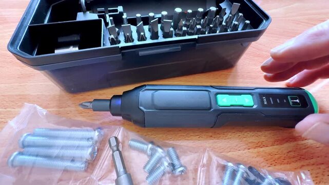 Closeup of a man&rsquo;s hand testing the forward and reverse power motion of a cordless stick screwdriver, between a box of varied bits and packets of bolt screw fastener fittings.