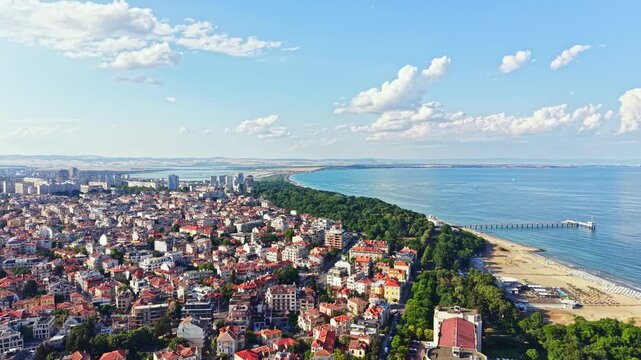 A view shows a city by the sea with buildings, trees, and a beach. The sky is clear with some clouds. People visit the shore and enjoy the water.