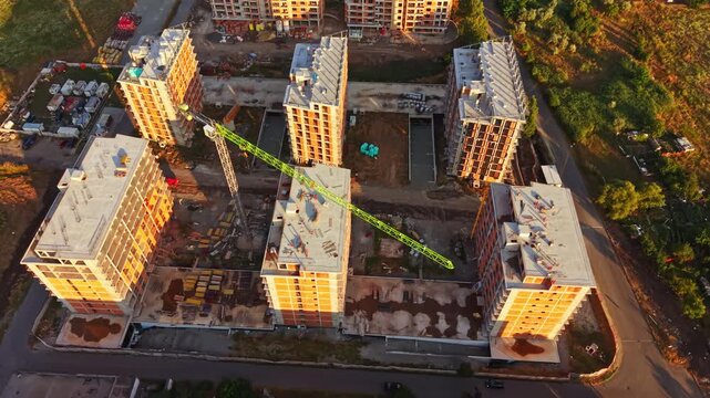 Workers are busy on a construction site with multiple buildings under development. A crane is moving materials around. The site shows ongoing work in the late afternoon sun.