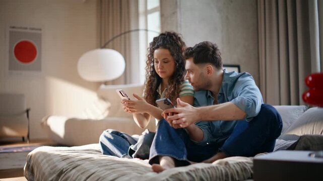 Couple using smartphones sitting bed home. Man reacting exciting news on phone