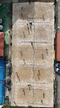 Imagem a&eacute;rea vertical de partida de v&ocirc;lei em quadra de areia, com atletas, rede central e linhas demarcadas, destacando esporte, movimento, integra&ccedil;&atilde;o e lazer ao ar livre.