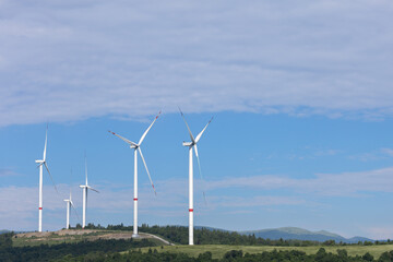 Fototapeta premium A view of multiple large white wind turbines on a green hill, with rolling mountains and a blue sky in the background with Copy space