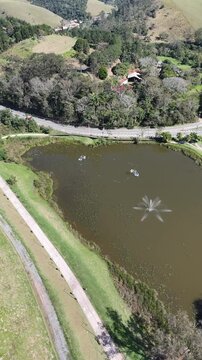 Imagem de drone do Parque do Lago, em Guararema, mostrando lago ornamental, chafariz, gramados, vias curvas e montanhas em cen&aacute;rio de lazer, turismo e natureza.
