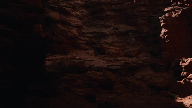 Visitors walk in a dark cave. Light shines on the rock walls, revealing textures and colors. The ground is uneven with scattered stones. Shadows create depth and mystery in the cave.