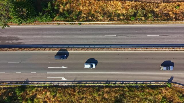 Cars and trucks drive on a highway surrounded by grassy areas. The sun is shining down on the road. Some vehicles move slower while others speed by.