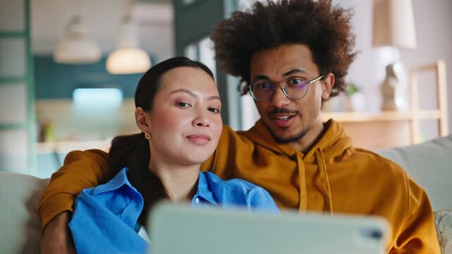 Multiracial couple browsing tablet on sofa at home. Pair relaxing together.