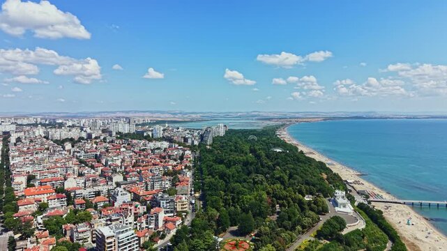 High-angle view of low-rise homes with tiled roofs, green parkland, broad sandy shoreline, and calm ocean stretching to horizon, creating scenic coastal city landscape with relaxing summer light