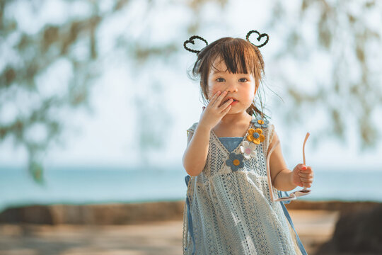 Cute little Asian girl holding sunglasses while standing on beach during summer vacation, portrait of happy toddler child looking at camera with sea background for family travel tourism, parenting.
