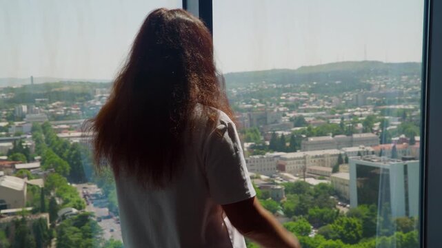 Silhouetted woman with long dark hair looks out floor to ceiling window from high rise apartment overlooking sprawling city, hills, buildings