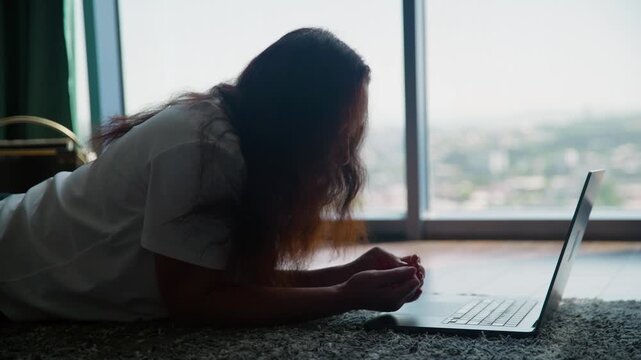 Woman reclined on carpet floor examining laptop screen with panoramic cityscape view through floor to ceiling windows behind her in high rise apartment.