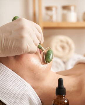 Gloved hand rolling jade facial roller across serum-glossed cheek skin in bright spa setting