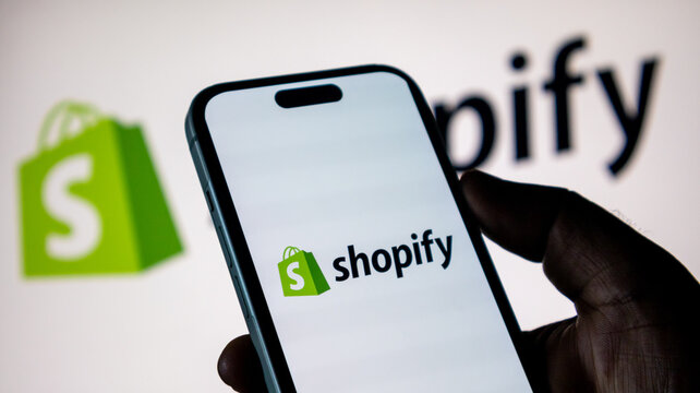 Hand holding smartphone with Shopify logo against blurred background screen.