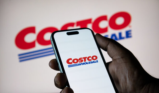 Man holding modern smartphone with Costco wholesale logo displayed on screen in front of blurred background.