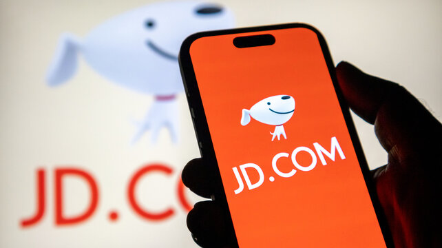 Human hand holding smartphone showing red JD dot com brand logo icon on display with blurred logo in background.