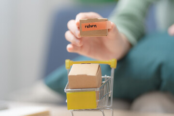 Online shopping return policy concept. Customer holding a parcel labeled return above a miniature shopping cart at home. Return, ecommerce, refund, exchange, delivery © Thitiporn