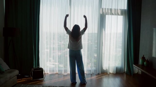 Silhouetted woman standing at floor to ceiling window with arms raised in celebration, overlooking expansive city skyline from luxury high rise apartment, backlit natural light through sheer curtains.