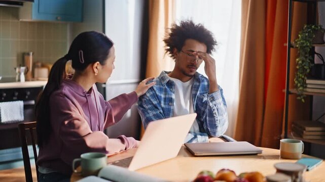 Multiracial couple stressed over bills at kitchen table, woman comforts man