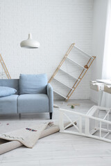 Messy living room with fallen furniture and scattered items, home disaster and renovation concept, a light blue sofa sits next to a toppled wooden and white bookshelf © Pixel-Shot