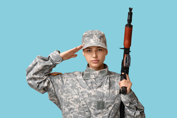 Fototapeta premium Confident young female soldier with assault rifle saluting on blue background